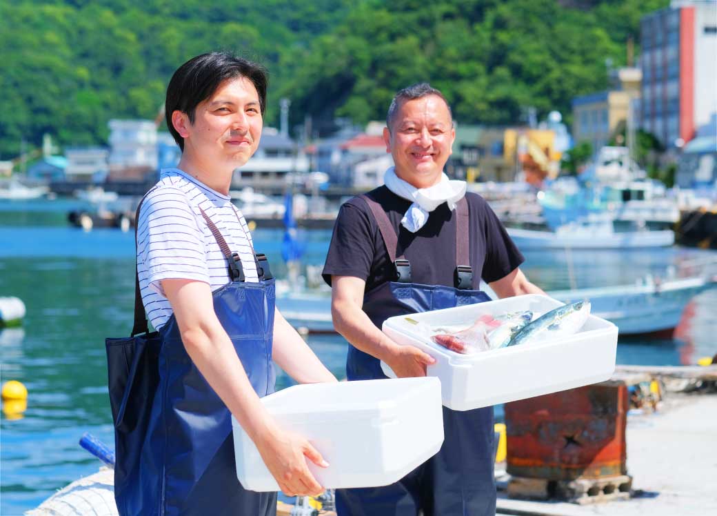 船の上で、魚などが入った容器を両手で持っている笑顔の男性2名